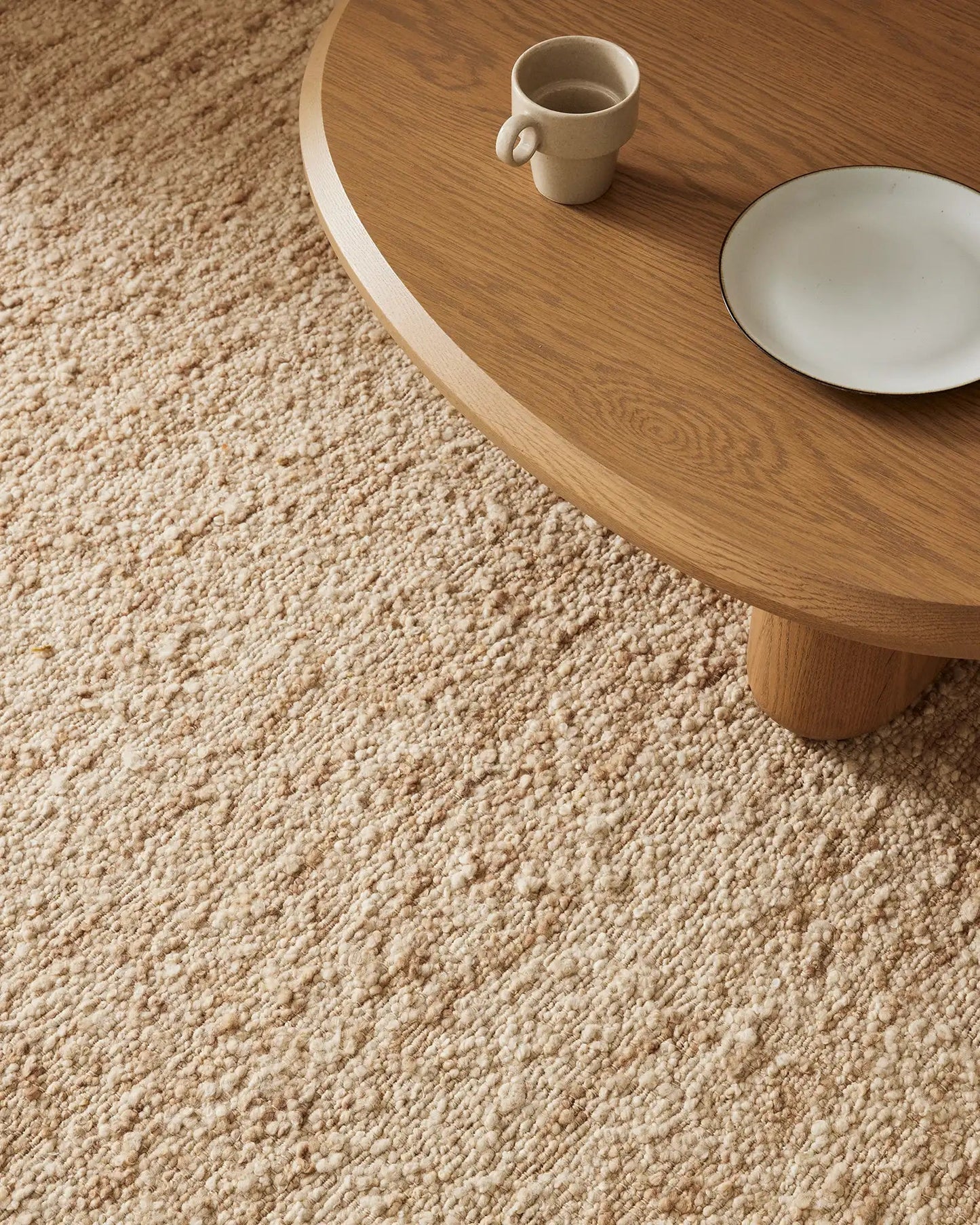 A close-up image of a beige Henley floor rug made from wool, featuring a textured chunky weave design, with a wooden table and chairs in the background.
